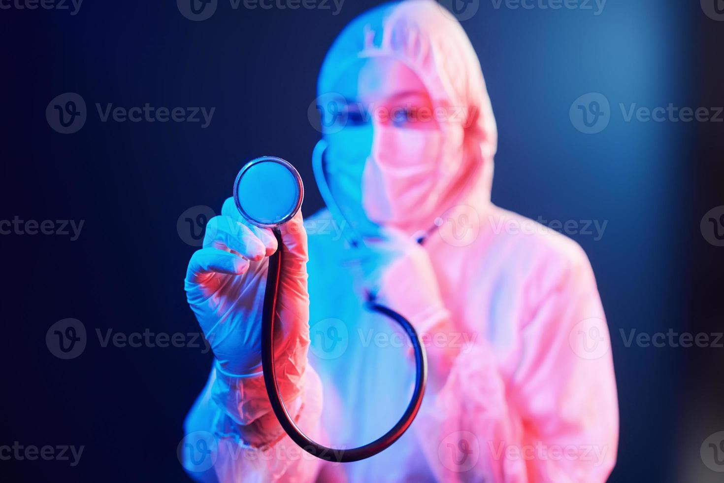 verpleegster in masker en wit uniform en met stethoscoop staand in neon verlicht kamer foto