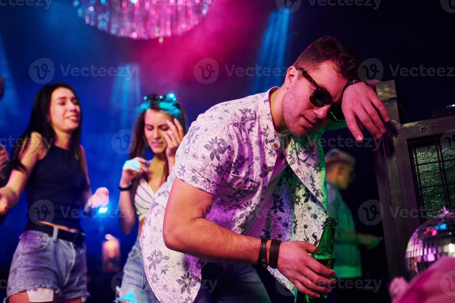 dronken vent leunend in hek in voorkant van jong mensen dat hebben pret in nacht club met kleurrijk laser lichten foto