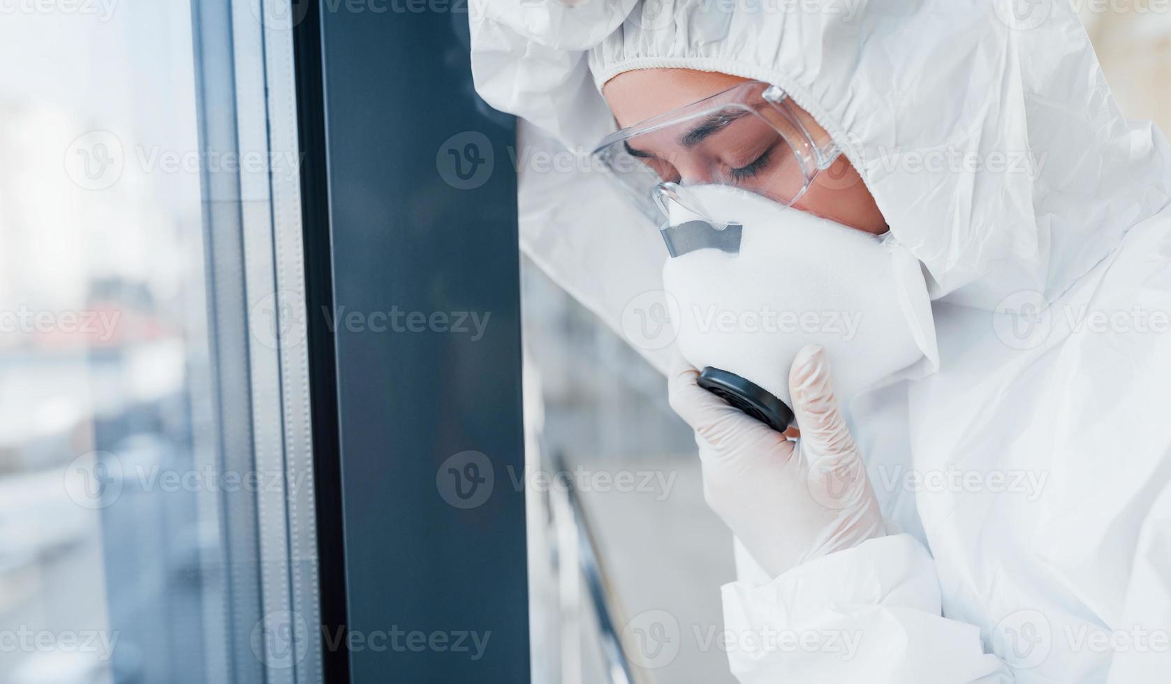 voelt slecht en ziek. vrouw dokter wetenschapper in laboratorium jas, defensief eyewear en masker staand binnenshuis foto