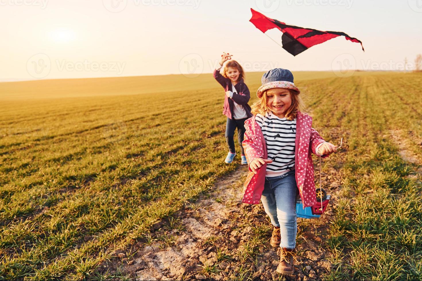 twee weinig meisjes vrienden hebben pret samen met vlieger en speelgoed- auto Aan de veld- Bij zonnig dag foto