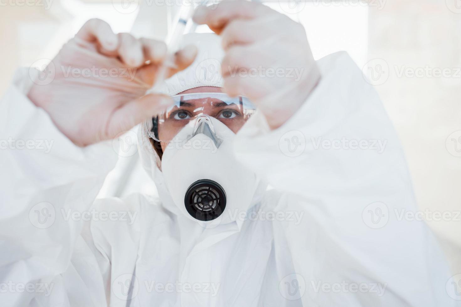 houdt injectiespuit met geneesmiddel. vrouw dokter wetenschapper in laboratorium jas, defensief eyewear en masker staand binnenshuis foto