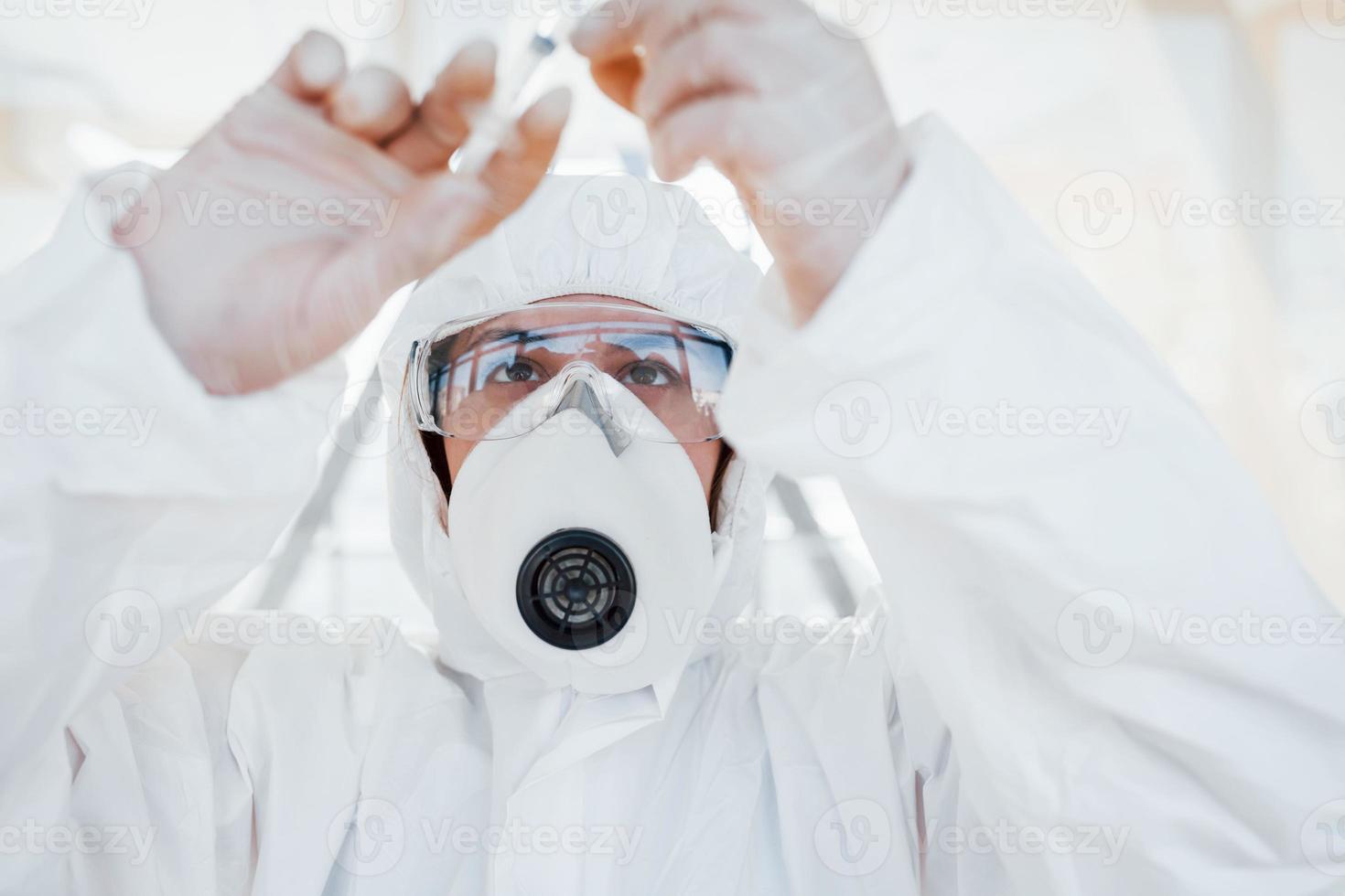 houdt injectiespuit met geneesmiddel. vrouw dokter wetenschapper in laboratorium jas, defensief eyewear en masker staand binnenshuis foto