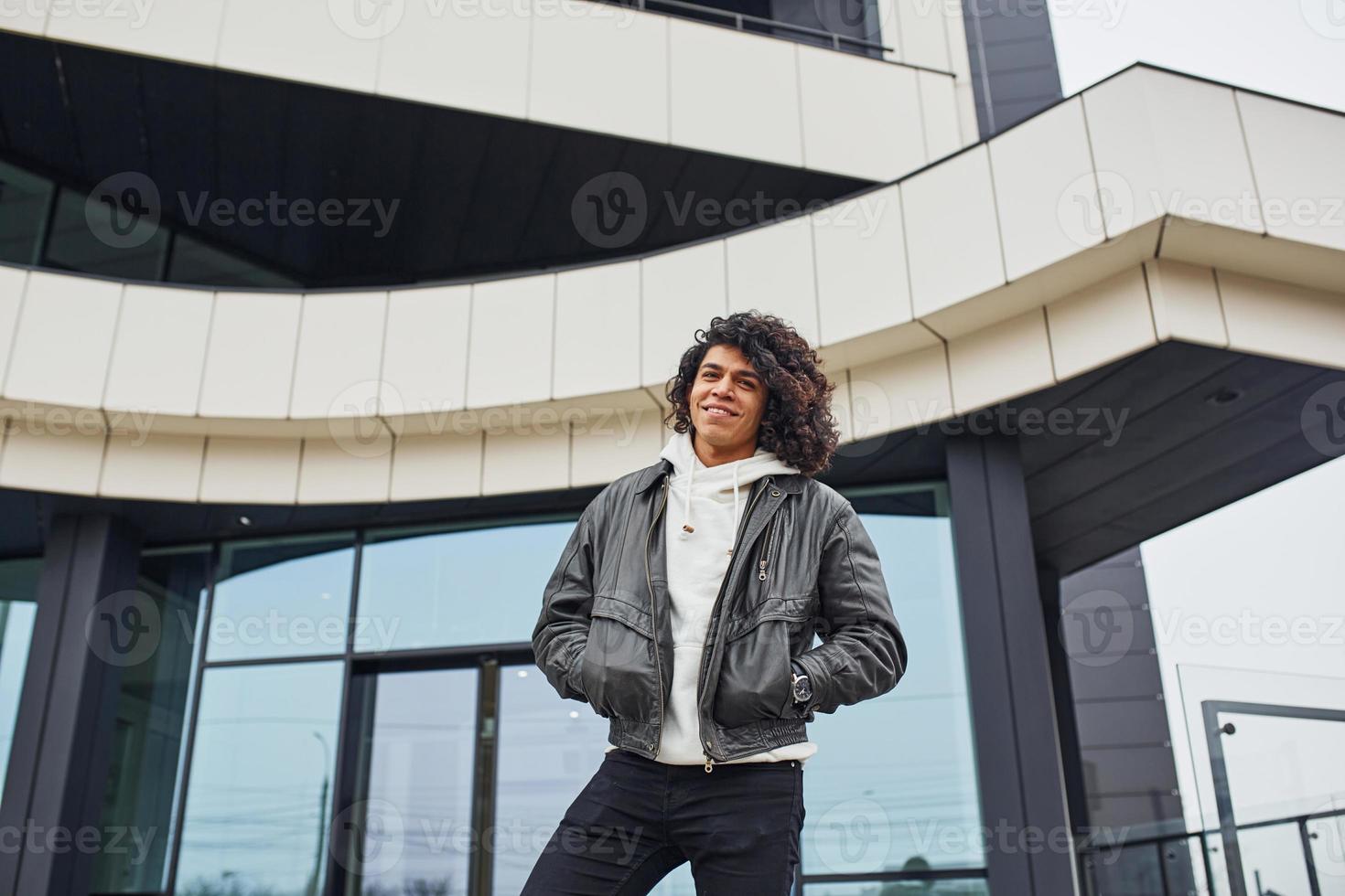 knap jong Mens met gekruld zwart haar- poseren voor de camera Aan de straat tegen gebouw foto