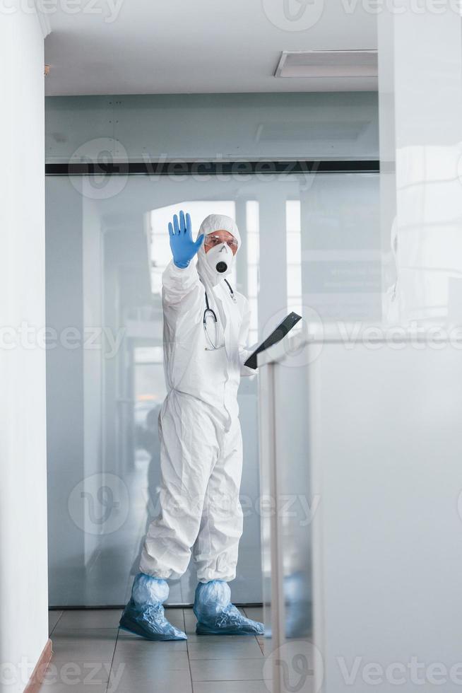 shows hou op gebaar door hand. mannetje dokter wetenschapper in laboratorium jas, defensief eyewear en masker Holding kladblok in handen foto