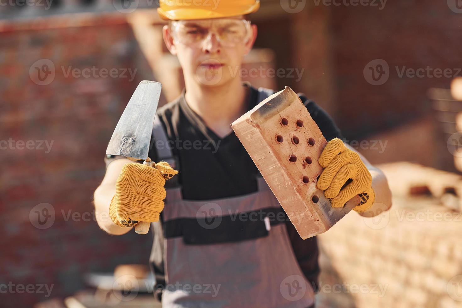 portret van bouw arbeider in uniform en veiligheid uitrusting dat staand Aan gebouw en Holding steen en gereedschap foto