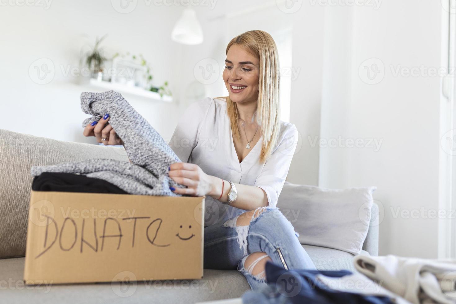 gelukkig blond jong vrouw zitten Aan bankstel zit vast kleren in bijdrage doos Bij huis, zorgzaam biraciaal vrouw vrijwilliger zetten kleding in karton pakket, schenken naar behoeftig mensen, hergebruik, recycle concept foto