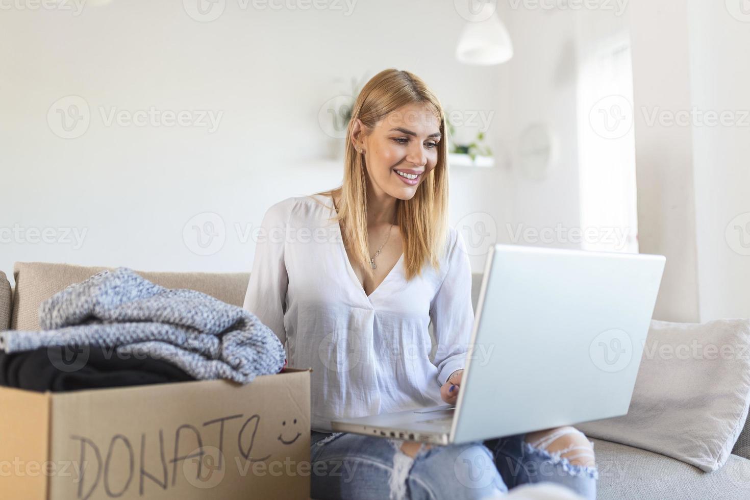 gelukkig jong vrouw zitten Aan bankstel zit vast kleren in bijdrage doos Bij huis, zorgzaam biraciaal vrouw vrijwilliger zetten kleding in karton pakket, schenken naar behoeftig mensen, hergebruik, recycle concept foto