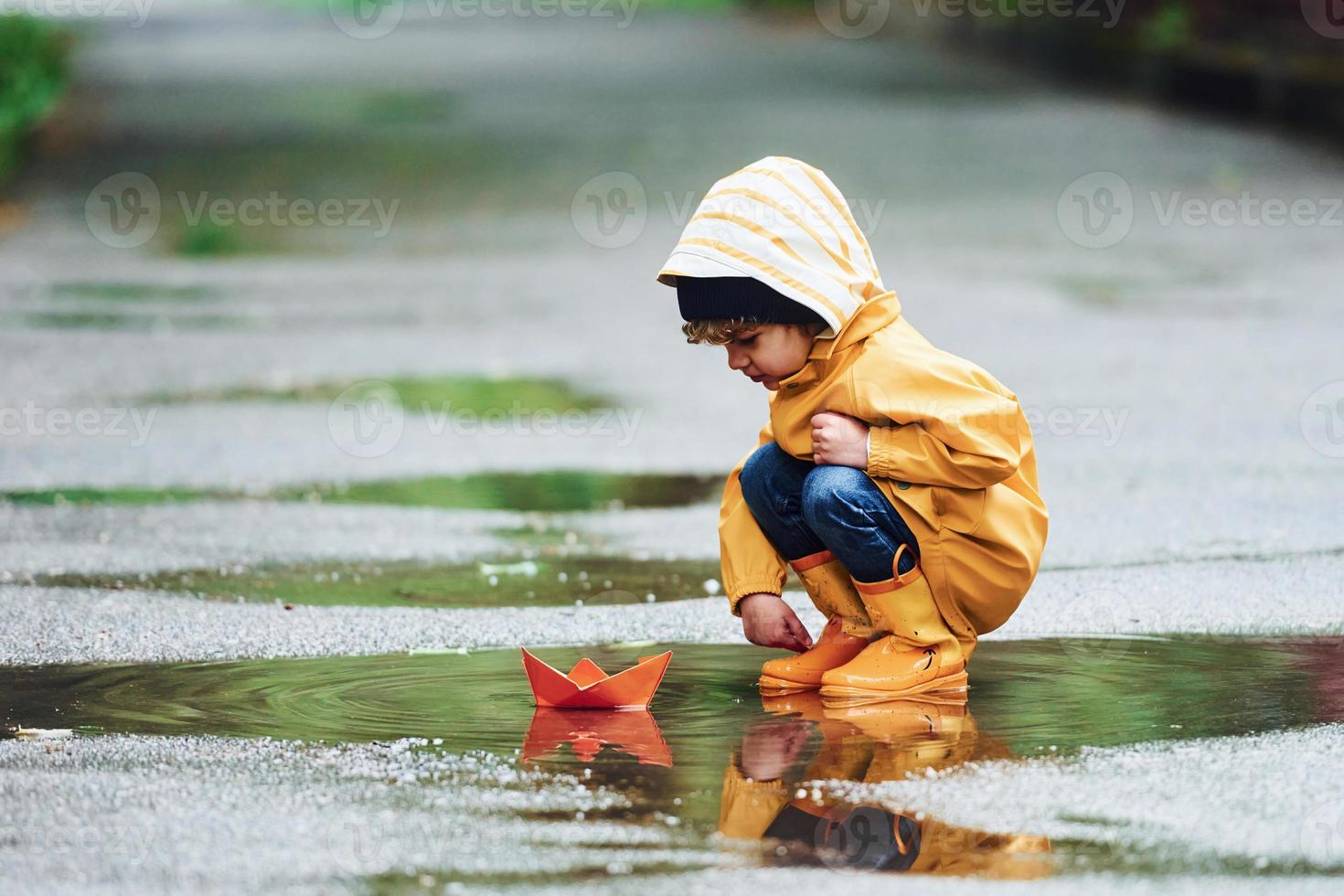 kind in geel waterbestendig mantel en laarzen spelen met papier handgemaakt boot speelgoed- buitenshuis na de regen foto