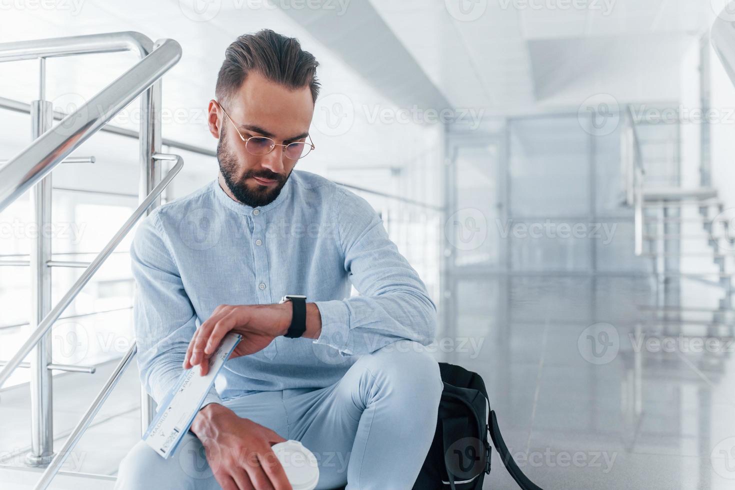 Holding vlak kaartjes in hand. zittend Aan trap en aan het wachten. jong passagier in formeel kleren binnenshuis in de kantoor Bij dag foto