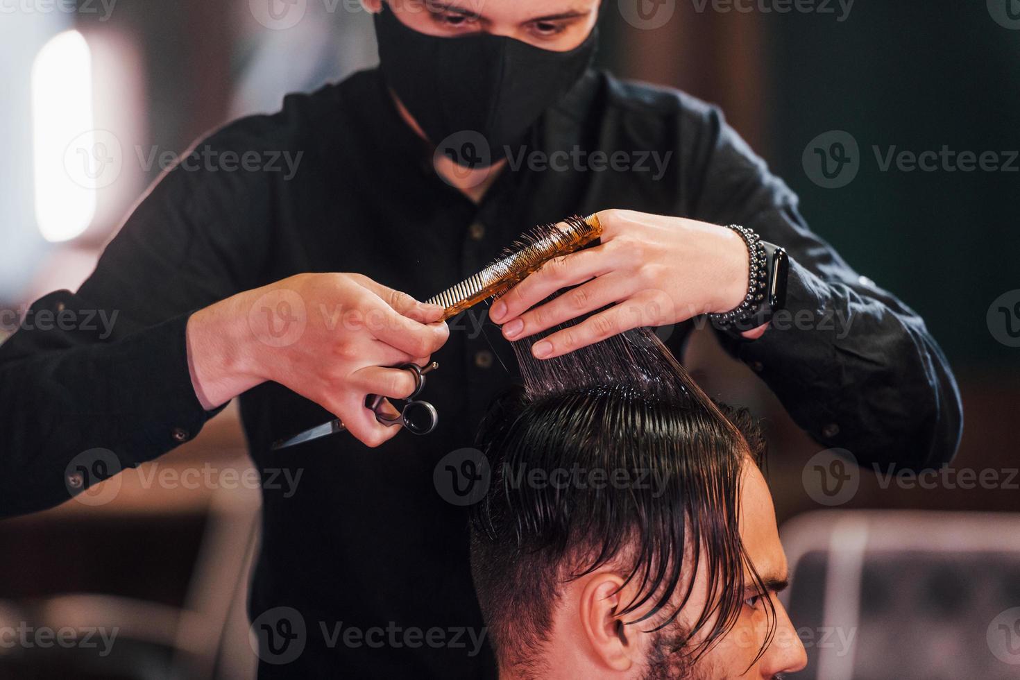 jong gebaard Mens zittend en krijgen kapsel in kapper winkel door vent in zwart beschermend masker foto