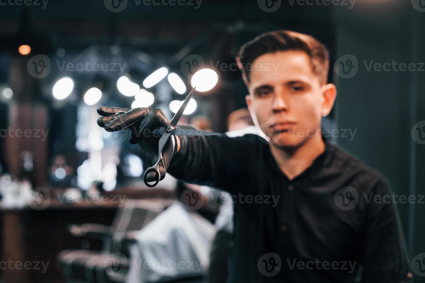 portret van jong vent in zwart kleren dat staand binnenshuis in kapper winkel en Holding schaar foto