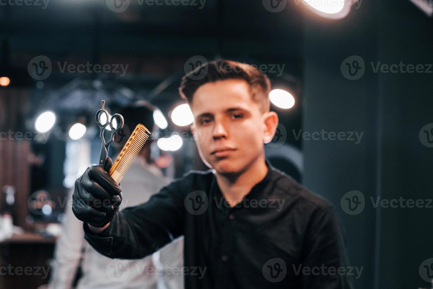 portret van jong vent in zwart kleren dat staand binnenshuis in kapper winkel en Holding schaar en kam foto