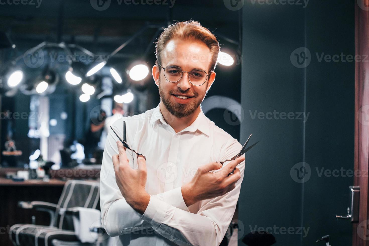 portret van jong vent in wit kleren dat staand binnenshuis in kapper winkel en Holding schaar foto