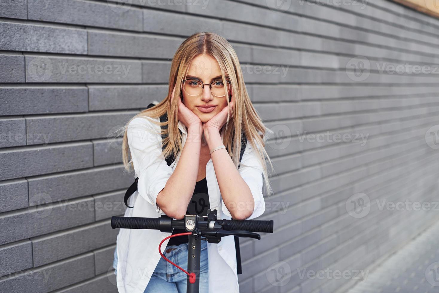 tegen grijs muur. mooi blond in gewoontjes kleren rijden elektrisch schieter buitenshuis Bij zonnig dag foto