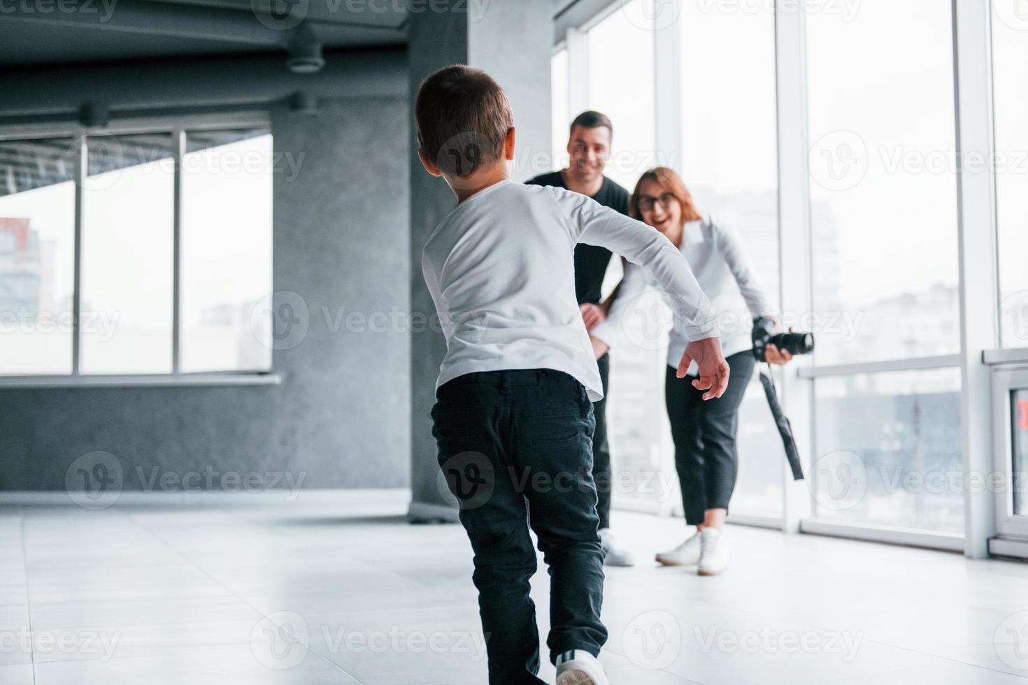 vrouw in formeel kleren en met camera in hand- staand binnen van leeg kamer met Mens en weinig jongen dat rennen en hebben pret foto