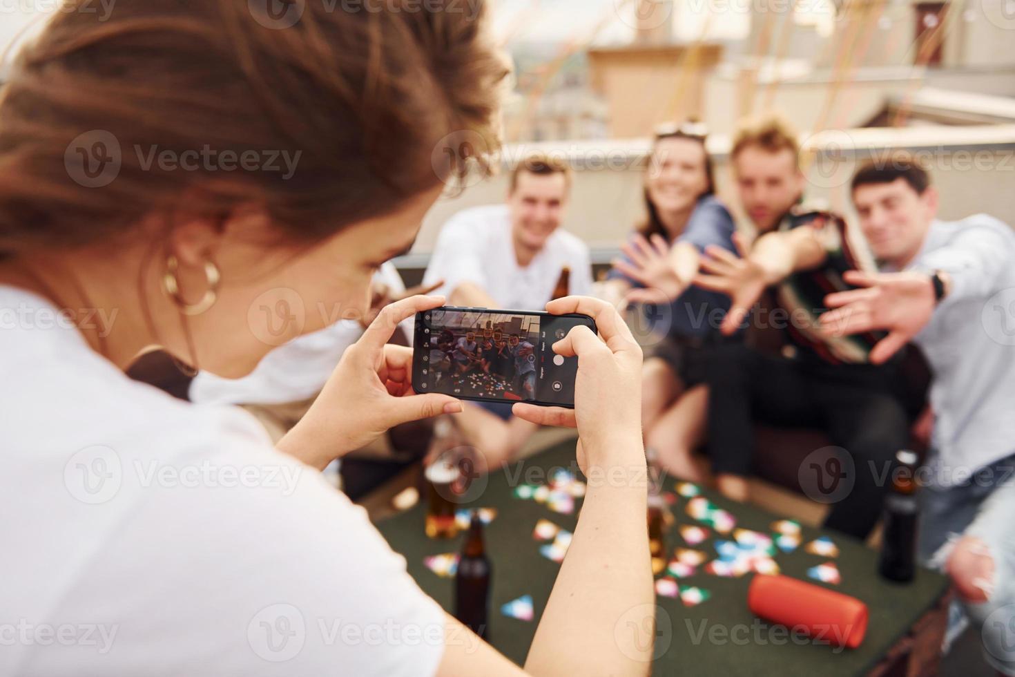 meisje aan het doen foto wanneer mensen spelen kaart spel. groep van jong mensen in gewoontjes kleren hebben een partij Bij op het dak samen Bij dag