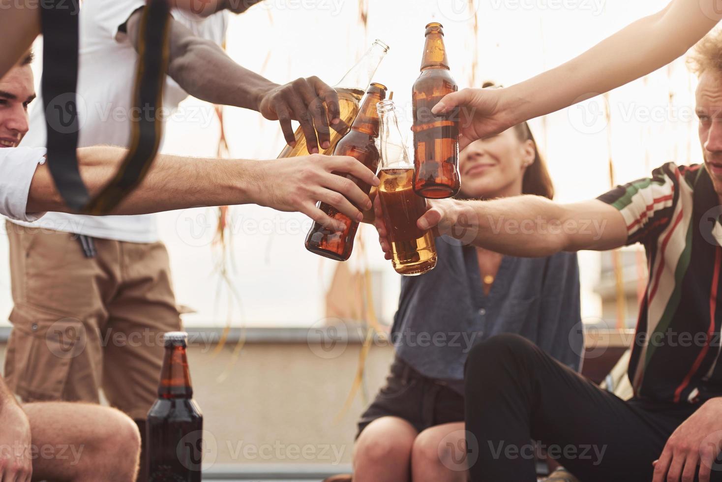 aan het doen proost door flessen van bier. groep van jong mensen in gewoontjes kleren hebben een partij Bij op het dak samen Bij dag foto