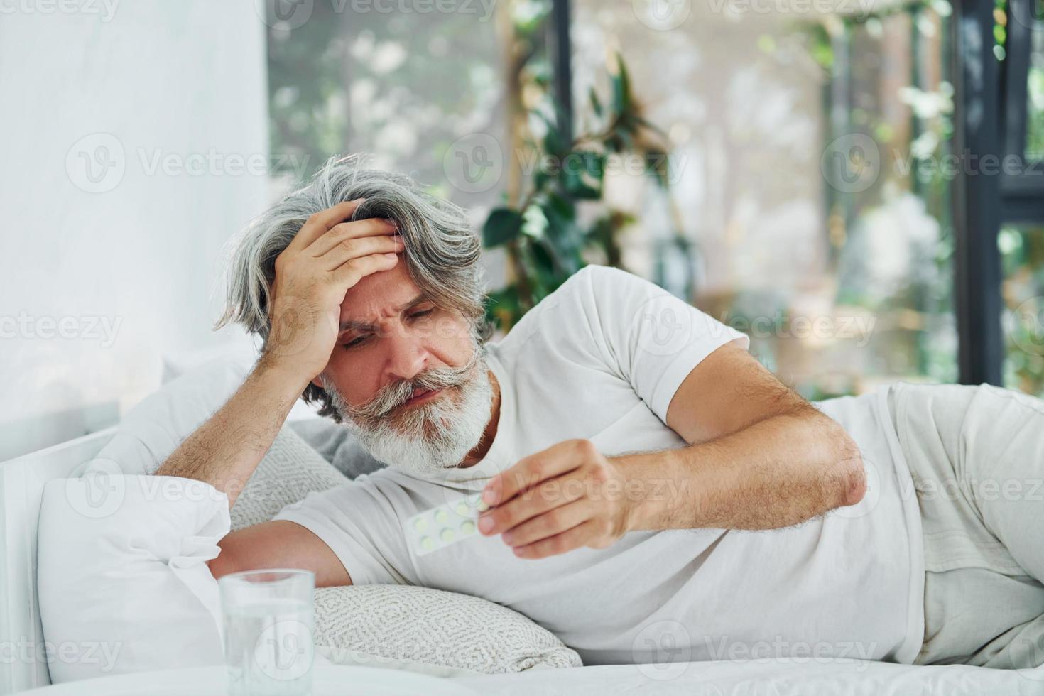 ziek senior elegant modern Mens met grijs haar- en baard binnenshuis aan het liegen Aan de bed en voelt slecht foto