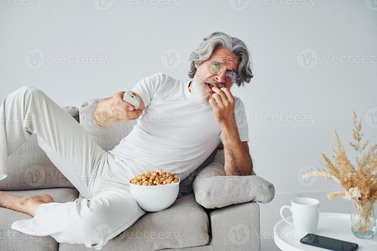 aan het kijken TV tonen en eet popcorn. senior elegant modern Mens met grijs haar- en baard binnenshuis foto