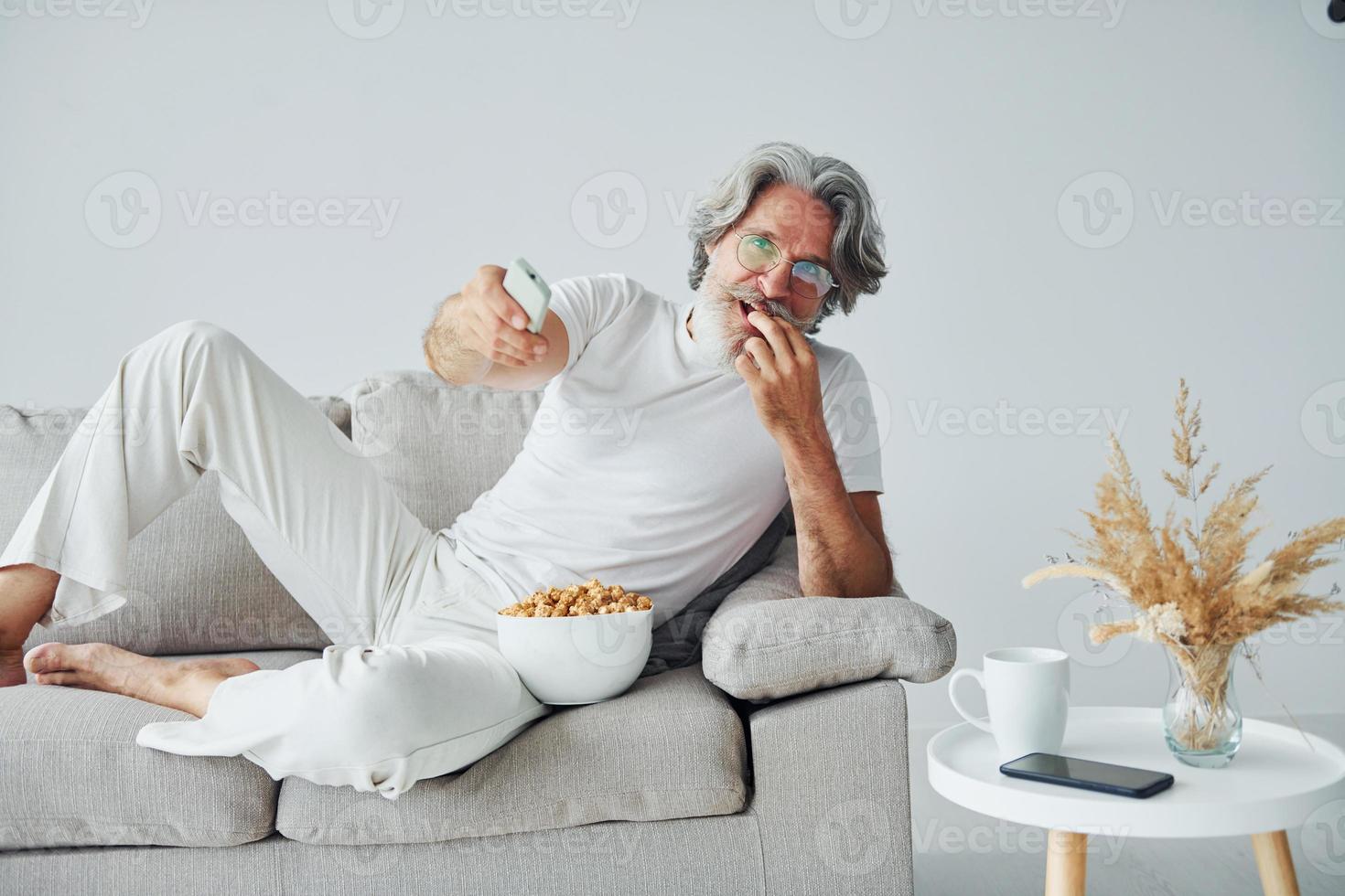 aan het kijken TV tonen en eet popcorn. senior elegant modern Mens met grijs haar- en baard binnenshuis foto