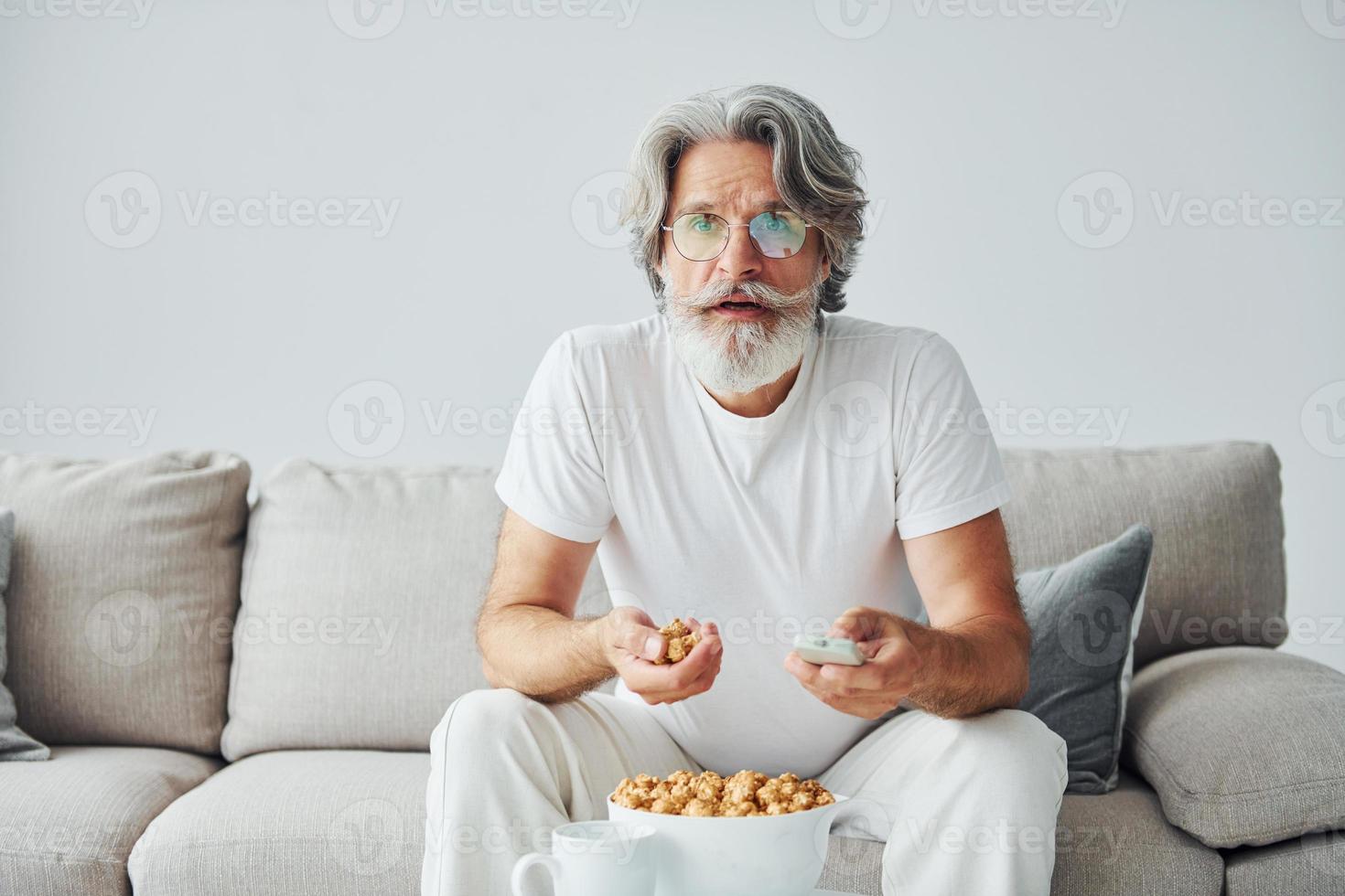 aan het kijken TV tonen en eet popcorn. senior elegant modern Mens met grijs haar- en baard binnenshuis foto