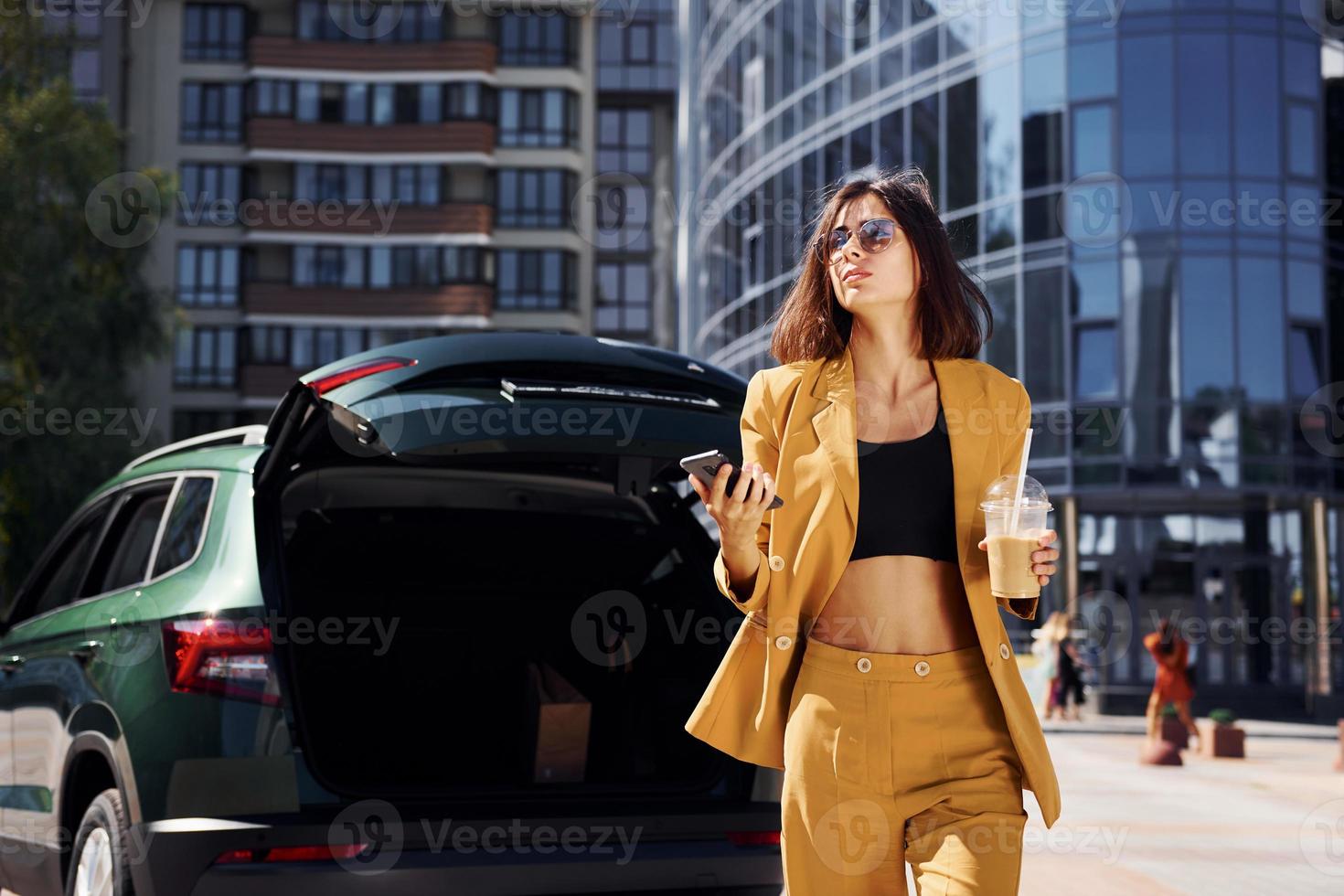 modern bedrijf gebouw Bij achtergrond. jong modieus vrouw in bordeaux gekleurde jas Bij dag met haar auto foto