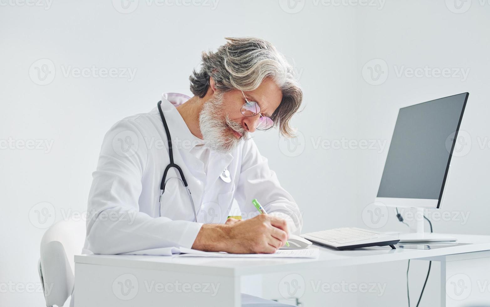 schrijft door computer tafel. senior mannetje dokter met grijs haar- en baard in wit jas is binnenshuis in kliniek foto
