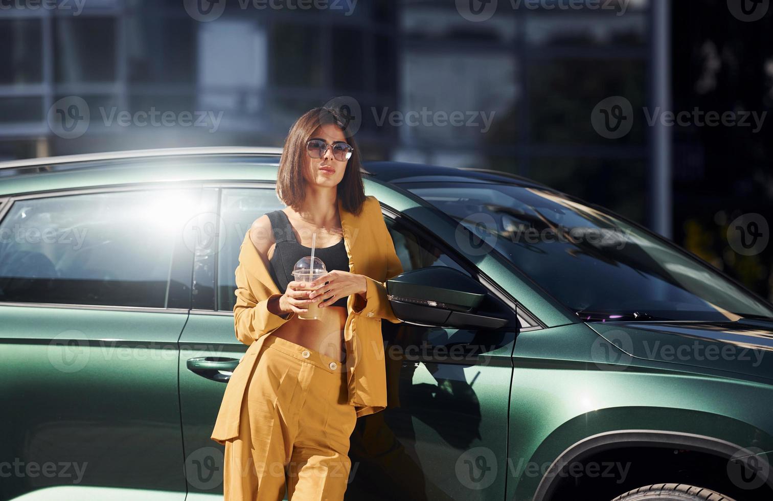met drinken in handen. jong modieus vrouw in bordeaux gekleurde jas Bij dag met haar auto foto