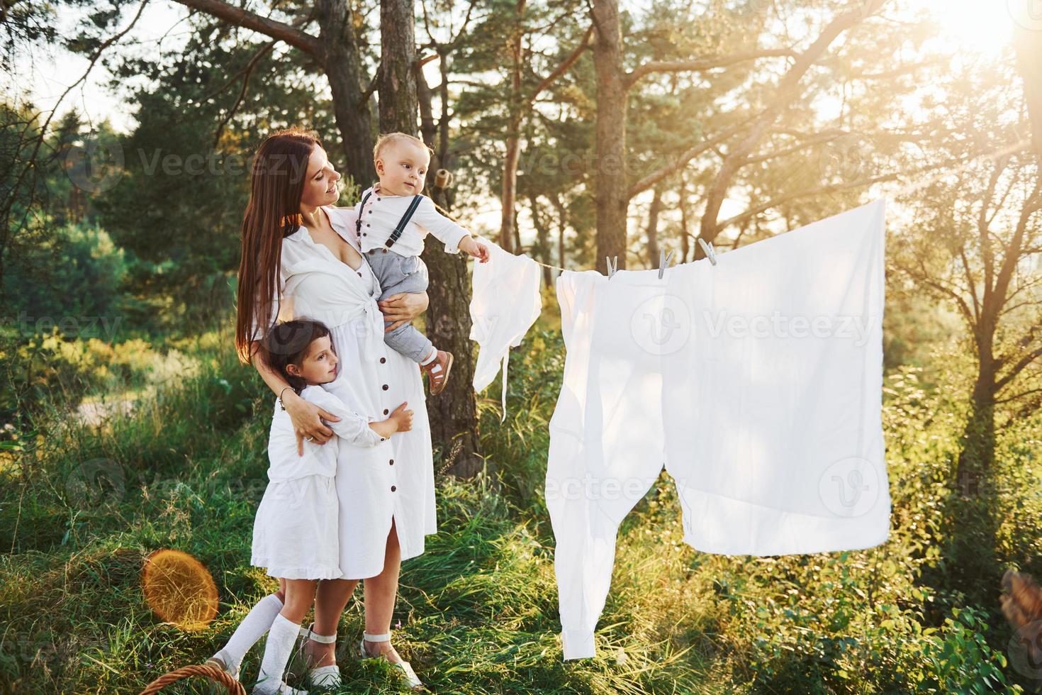 wit kleren hangende Aan de touw naar droog. jong moeder met haar weinig dochter en zoon is buitenshuis in de Woud. mooi zonneschijn foto