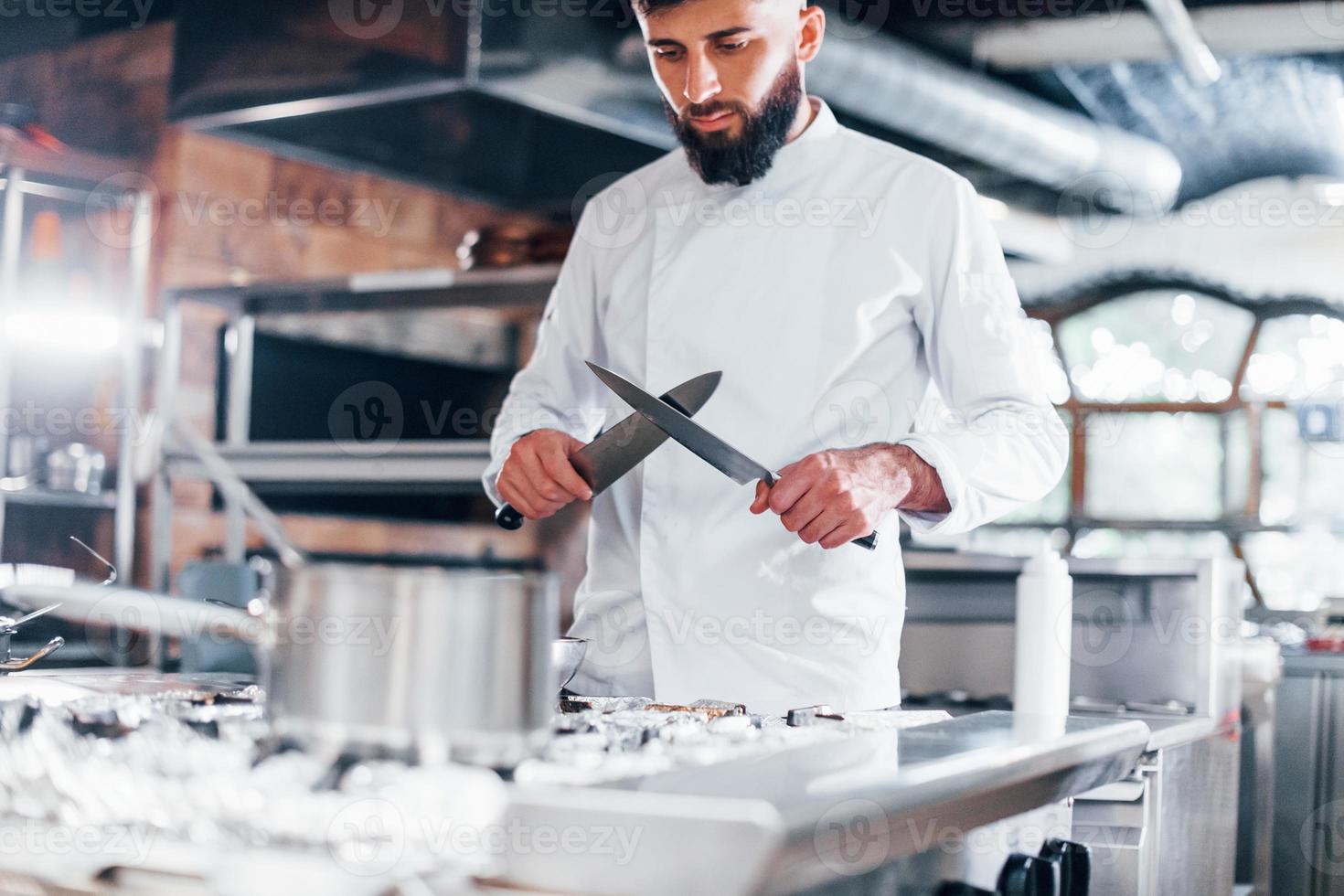 chef in wit uniform staand Bij keuken. Holding messen in handen foto