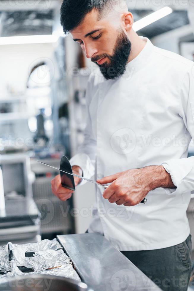 chef in wit uniform staand Bij keuken. Holding messen in handen foto