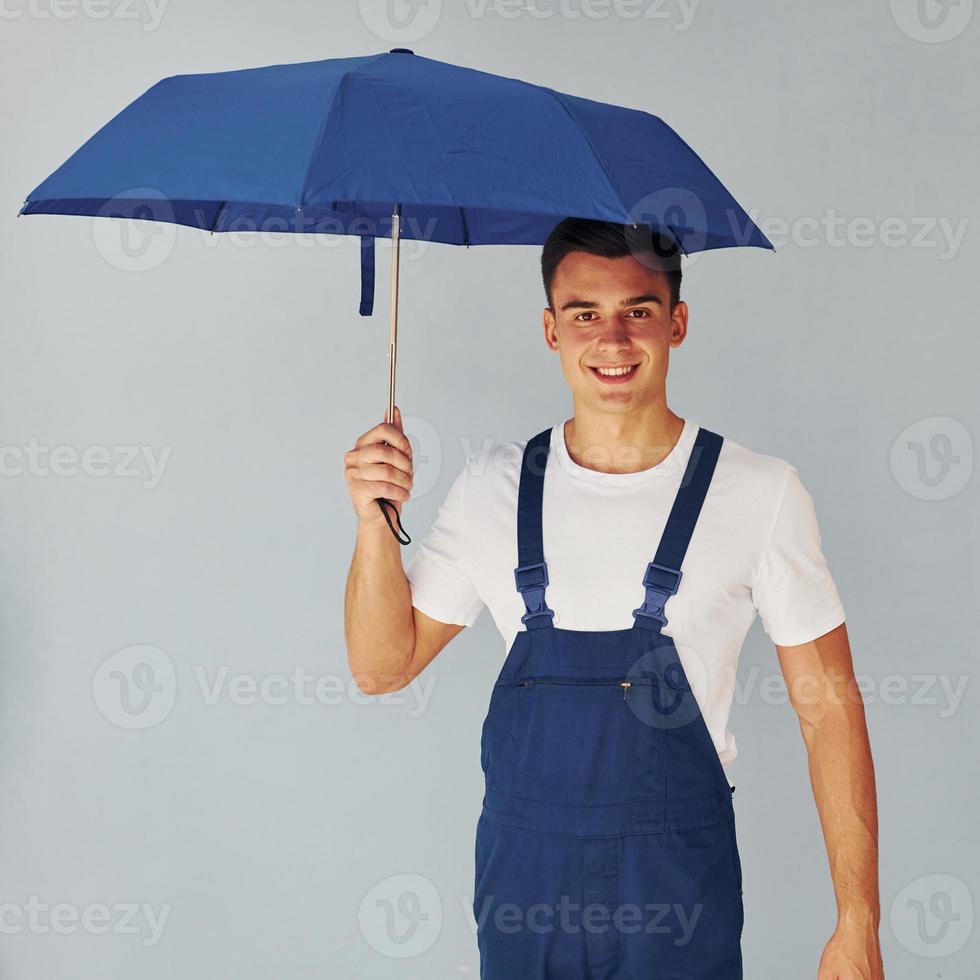 houdt paraplu door hand. mannetje arbeider in blauw uniform staand binnen van studio tegen wit achtergrond foto