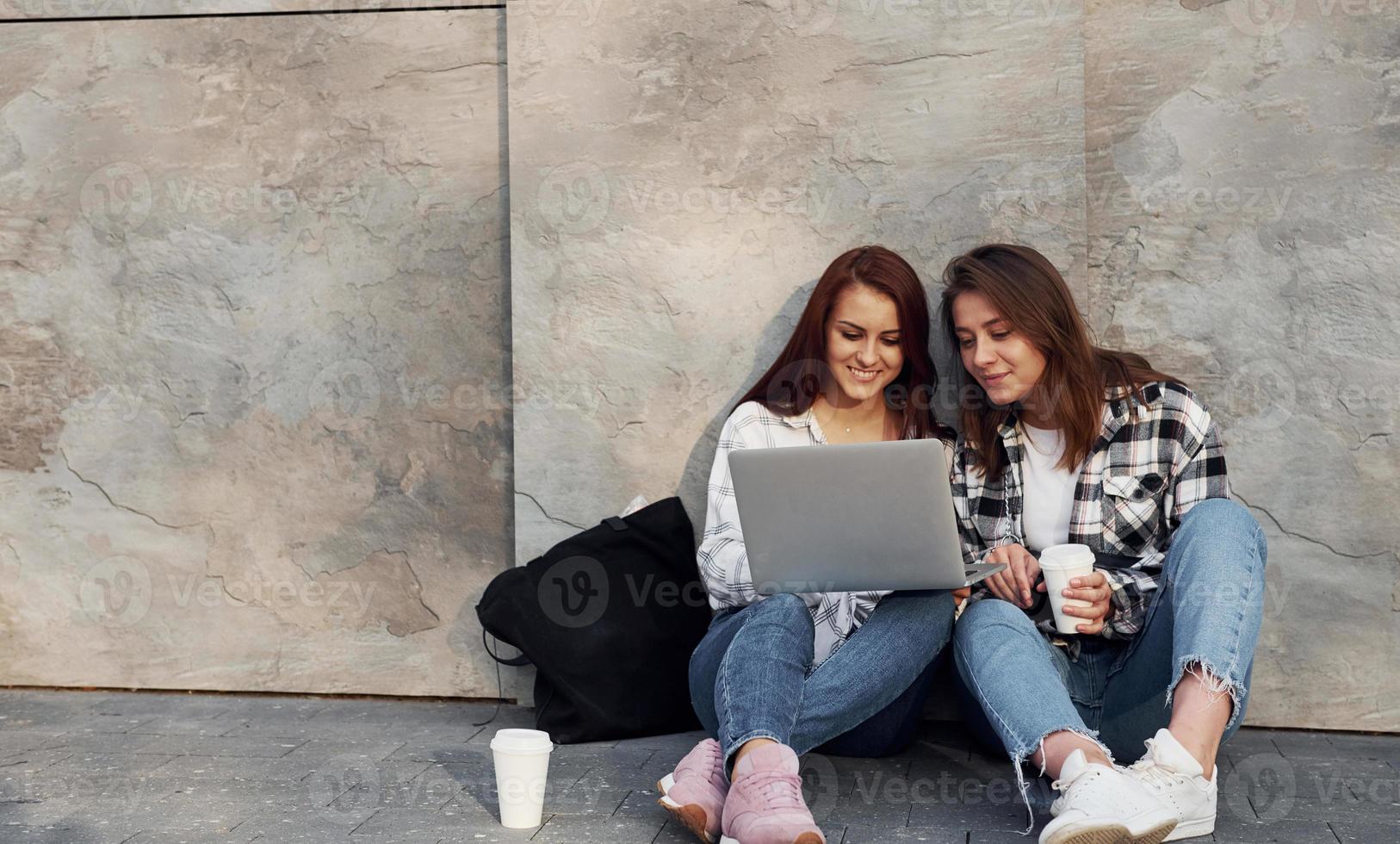 gebruik makend van laptop. mooi vrolijk vrienden of lesbienne paar samen in de buurt muur buitenshuis Bij dag foto