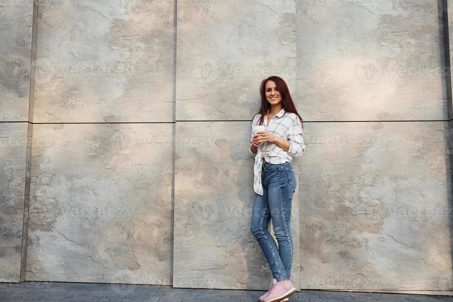 positief jong vrouw in gewoontjes kleren staand buitenshuis in de buurt muur met kop van drinken in handen foto