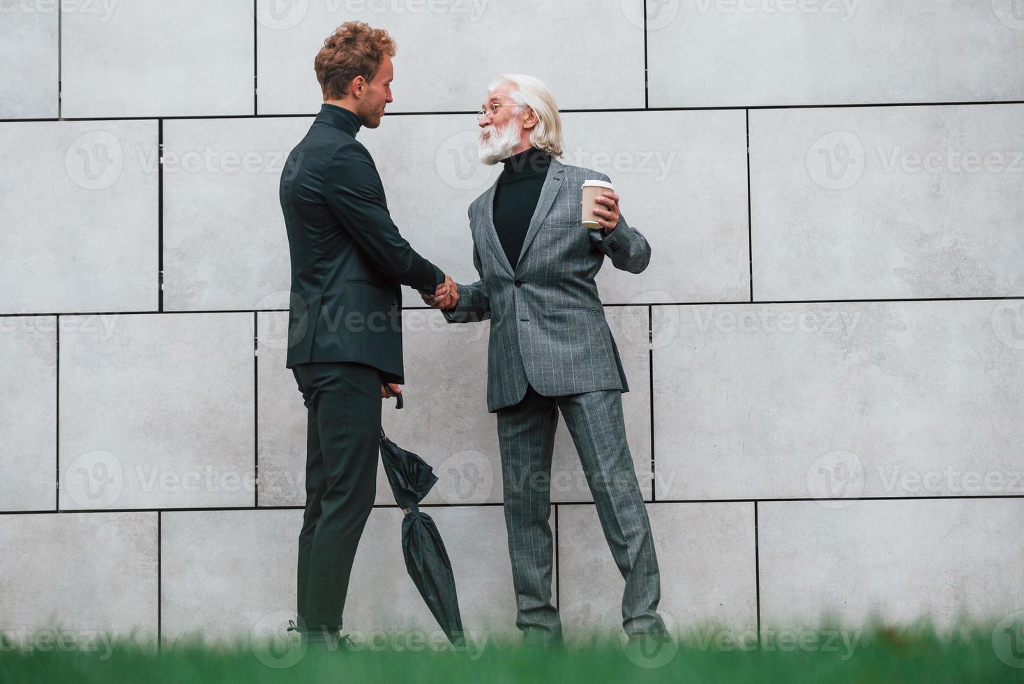 paraplu en kop van drankje. jong vent met senior Mens in elegant kleren is buitenshuis samen. opvatting van bedrijf foto