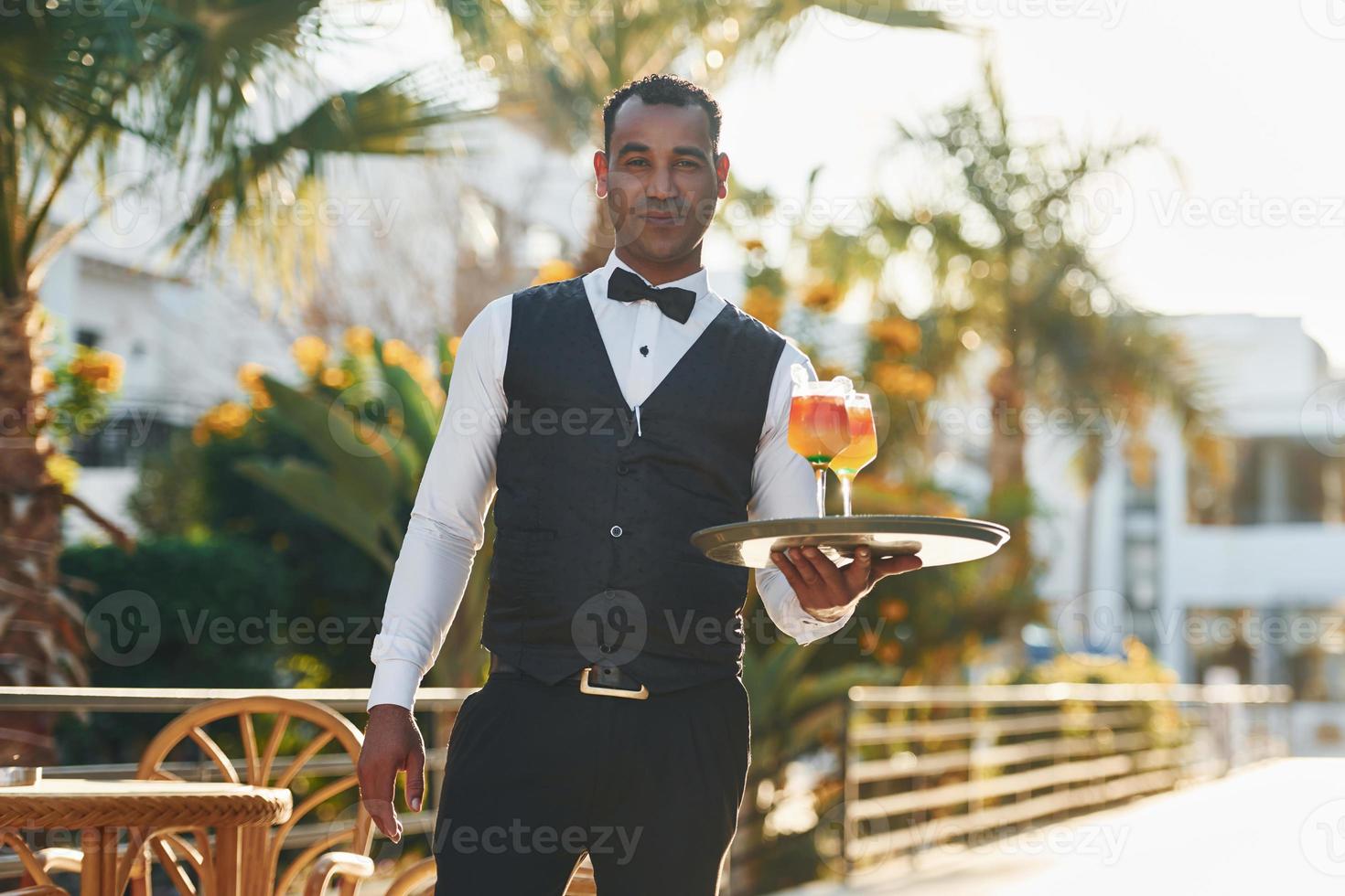 houdt cocktails. zwart ober in formeel kleren is Bij zijn werk buitenshuis Bij zonnig dag foto