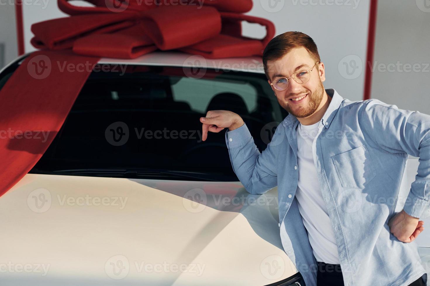 auto- is gedekt in geschenk plakband. geslaagd Mens in bril staand in de buurt merk nieuw auto binnenshuis foto