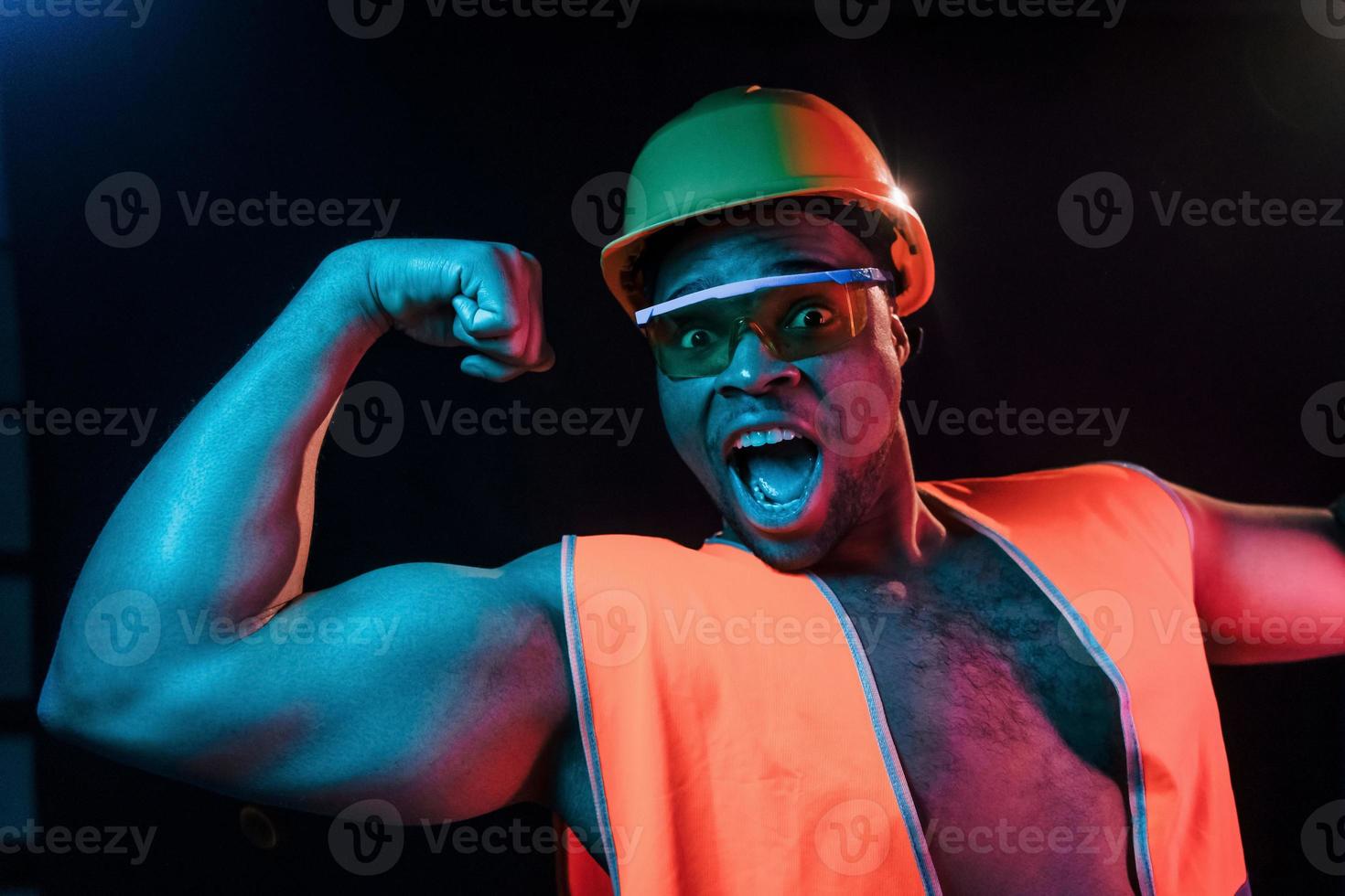 bouw arbeider in uniform en moeilijk hoed. futuristische neon verlichting. jong Afrikaanse Amerikaans Mens in de studio foto