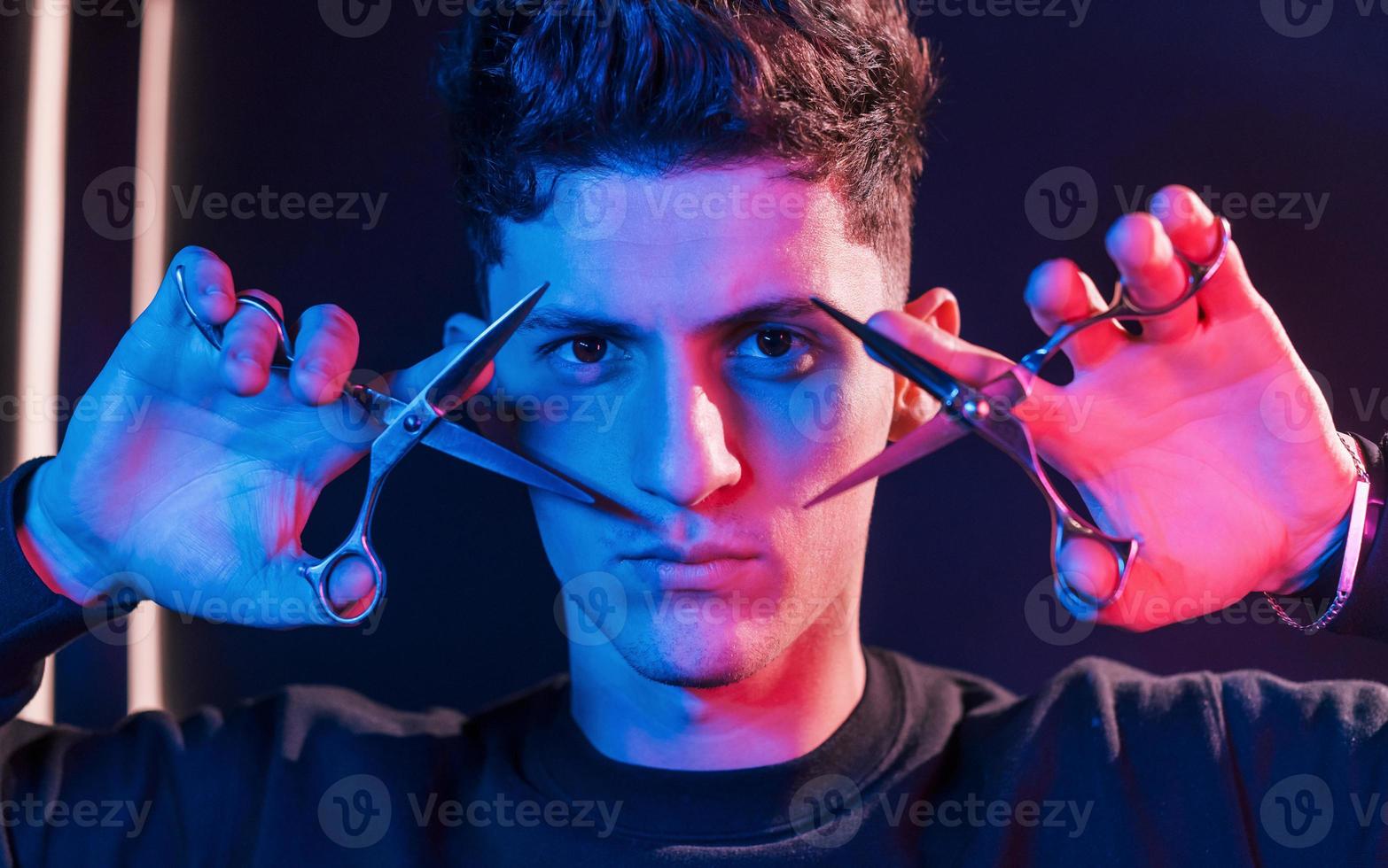 houdt schaar. jong kapper met werk uitrusting staand in de studio met neon verlichting foto