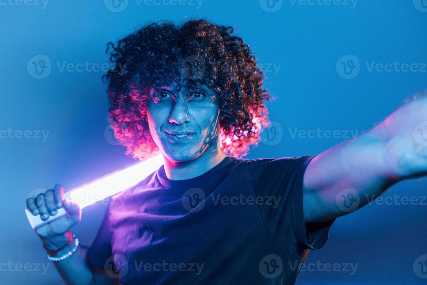 houdt verlichting apparatuur. jong mooi Mens met gekruld haar- is binnenshuis in de studio met neon verlichting foto