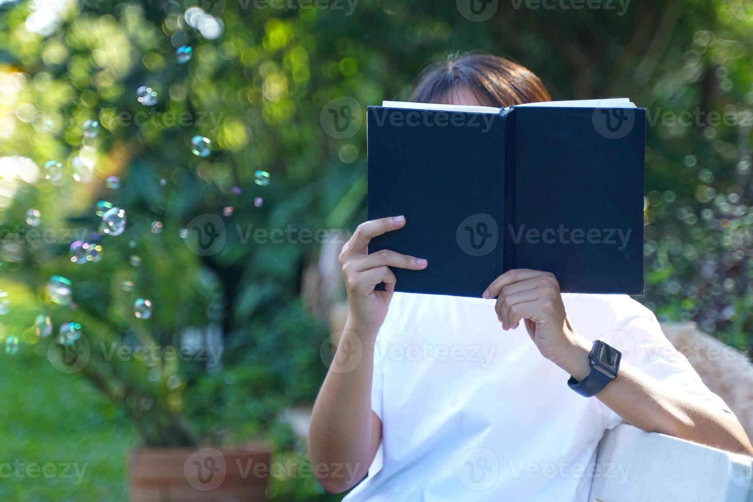 Aziatisch vrouw Holding een boek in voorkant van haar gezicht terwijl zittend en lezing park bank hoek. concept. Aziatisch vrouw aan het doen buitenshuis activiteiten, zo net zo lezing boeken, werken, hebben een picknick met familie foto