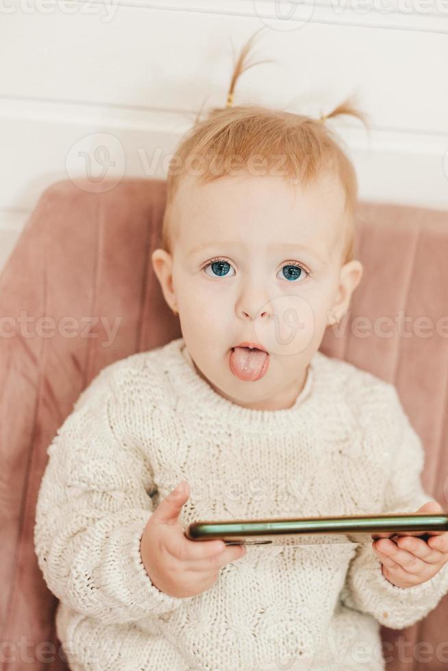 een weinig meisje met twee paardenstaarten stokjes uit haar tong en zit Aan een fauteuil met een telefoon in haar handen. grappig baby foto