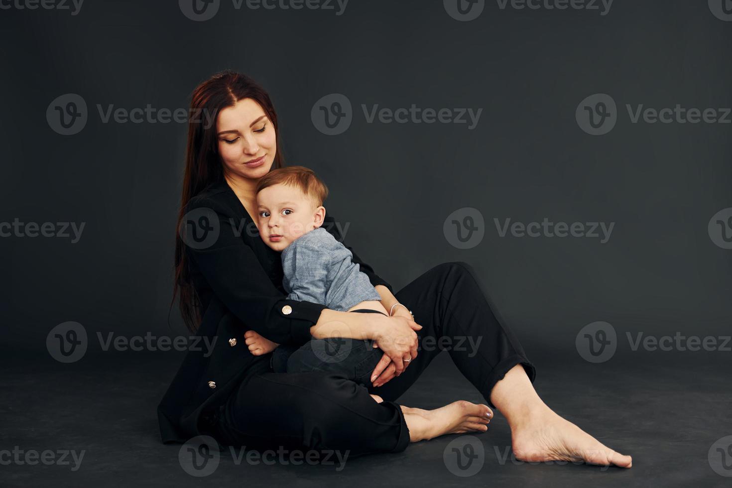 zittend Aan de vloer. moeder in elegant zwart kleren is met haar weinig zoon in de studio foto