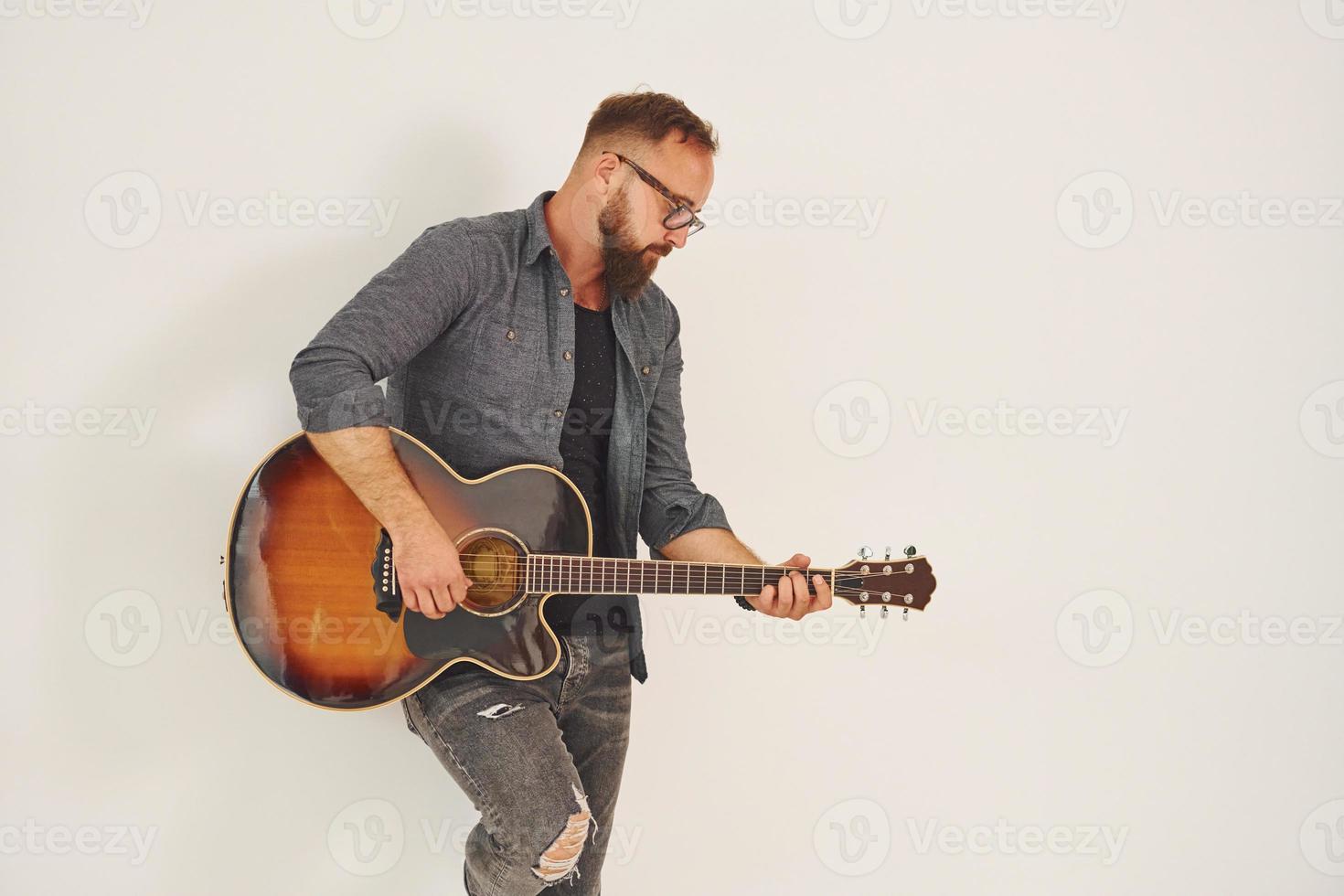 staand in de studio. Mens in gewoontjes kleren en met akoestisch gitaar is binnenshuis foto