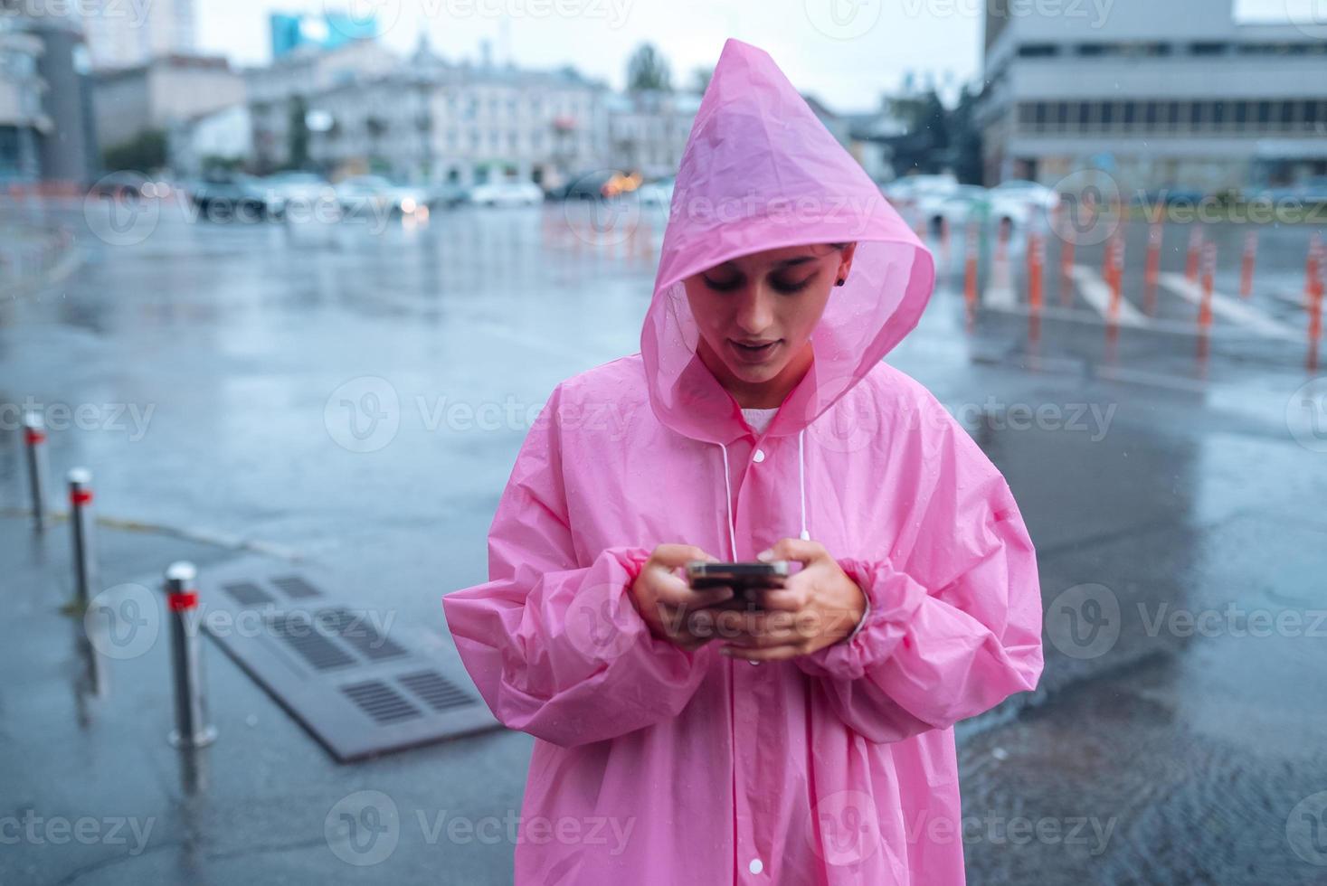 een jong vrouw in een regenjas op zoek Bij de smartphone foto