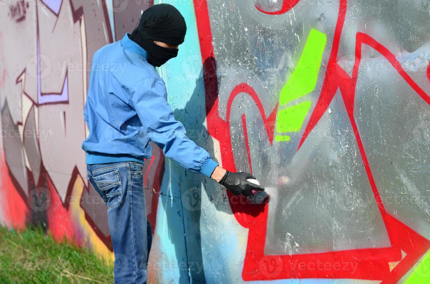 een jong hooligan met een verborgen gezicht verven graffiti Aan een metaal muur. onwettig vandalisme concept foto