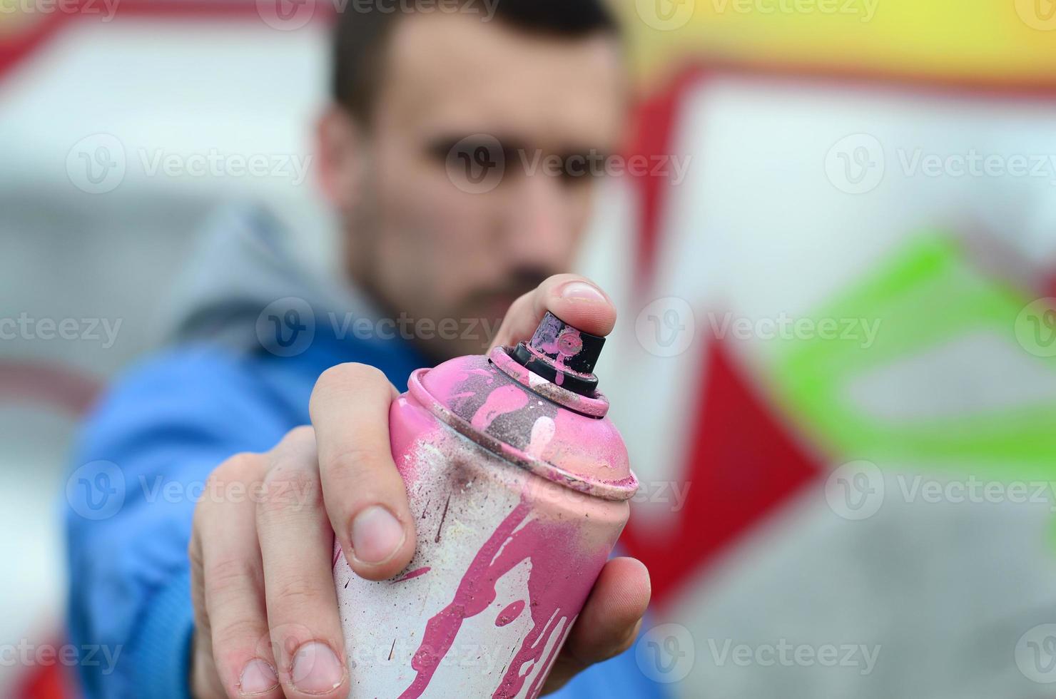 een jong graffiti artiest in een blauw jasje is Holding een kan van verf in voorkant van hem tegen een achtergrond van gekleurde graffiti tekening. straat kunst en vandalisme concept foto