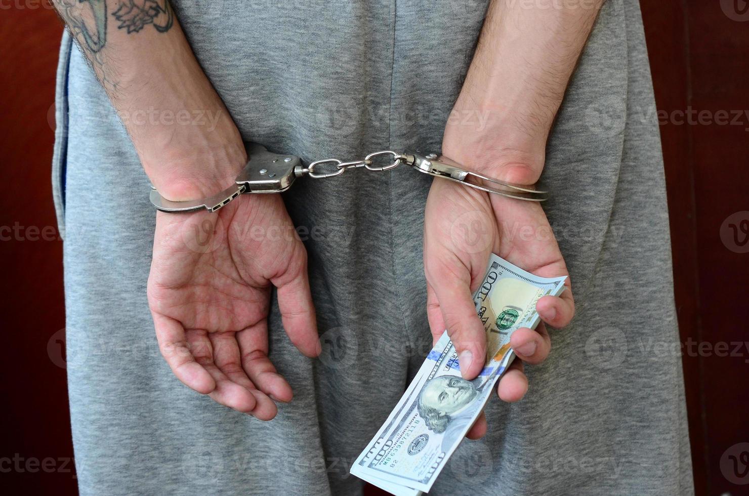 een gearresteerd vent in grijs broek met geboeid handen houdt een reusachtig bedrag van dollar rekeningen. terug visie foto