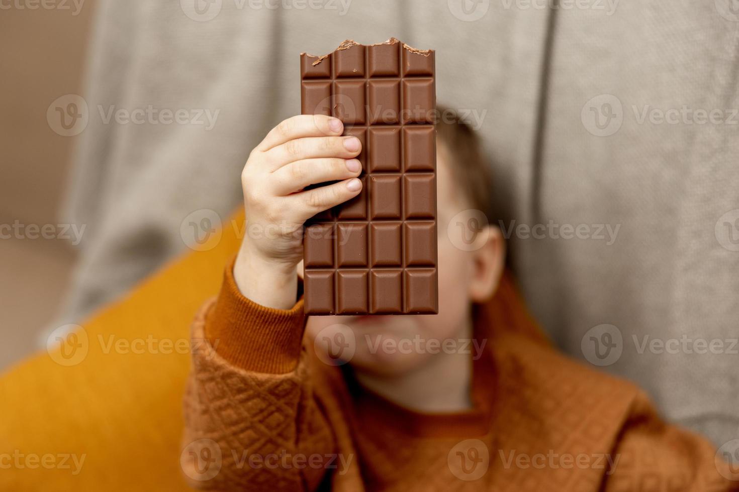 weinig aanbiddelijk jongen zittend Aan de bankstel Bij huis en aan het eten chocola bar. kind en snoepgoed, suiker banketbakkerij. kind genieten een heerlijk nagerecht. peuter- kind met gewoontjes kleding. foto