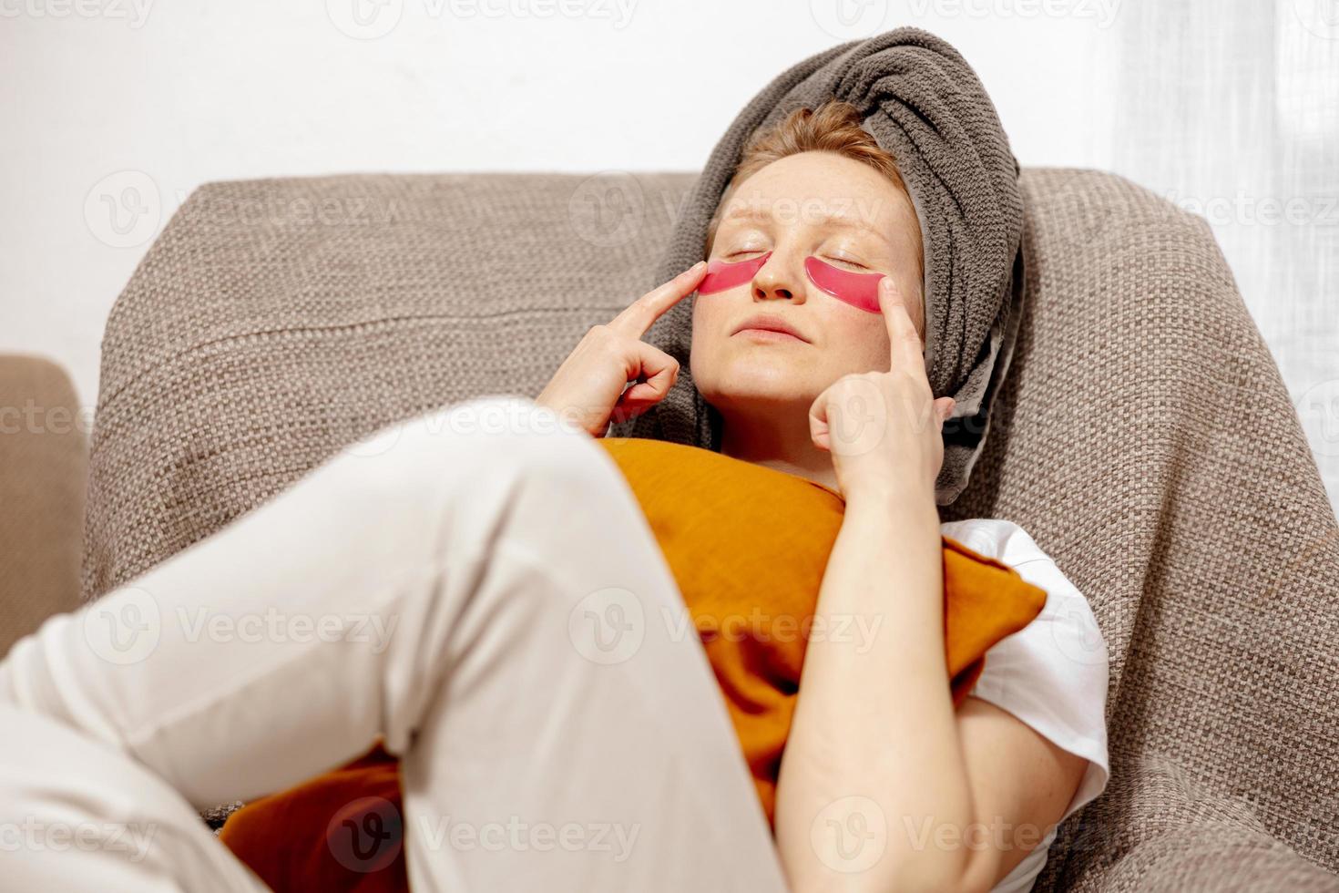 jong vrouw met gewoontjes kleren, handdoek Aan hoofd en oog patches zittend Aan bankstel en rusten. kom tot rust na nemen douche. kunstmatig voor Dames, huid zorg. zelf zorg, tijd voor jezelf. ontspannende Bij huis. foto